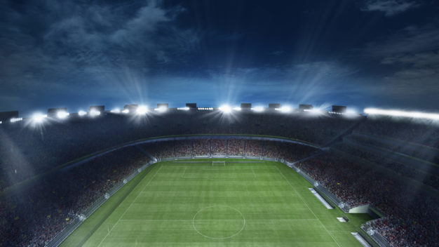 Empty football field with flashlights and dark night sky background. Stadium with filled stands with sports soccer fans.